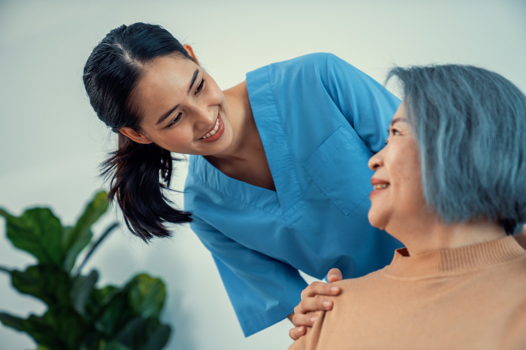 A caregiver rest her hands on the shoulders of a contented senior patient while she sitting on the sofa at home.